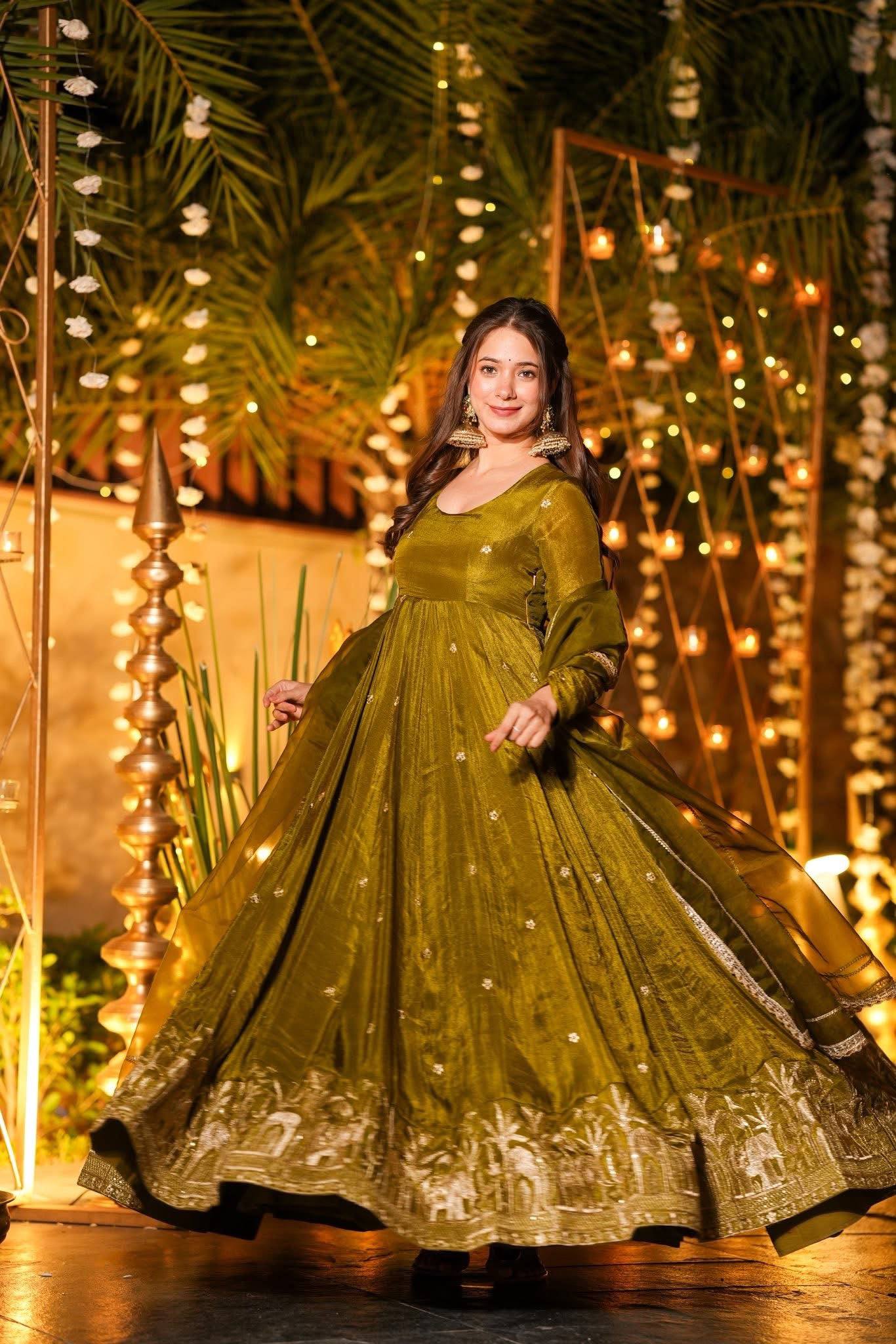 Woman in a green traditional outfit standing in front of decorative lights and plants.