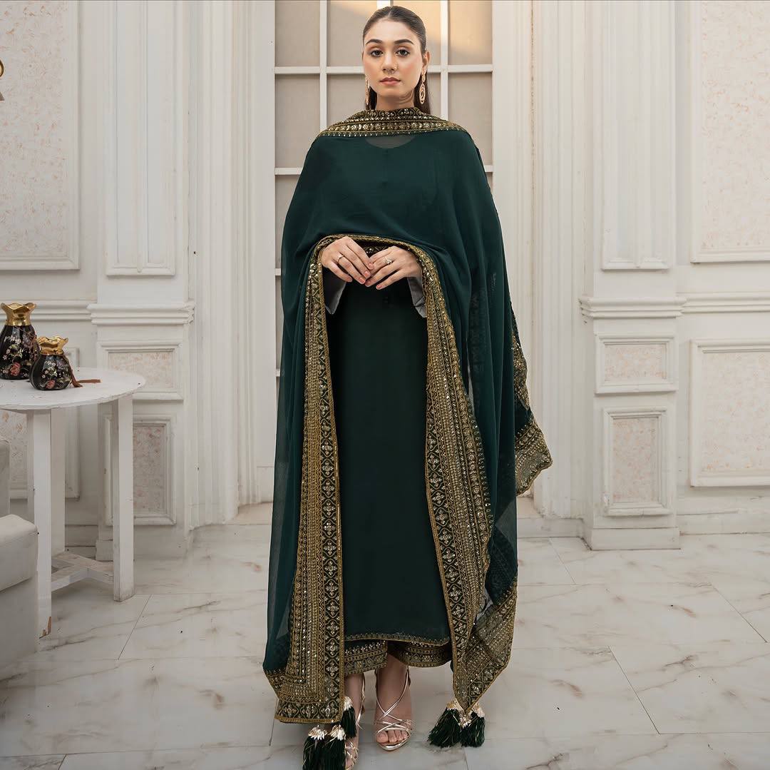 Woman in a dark green traditional outfit with gold embroidery standing in an elegant interior setting.