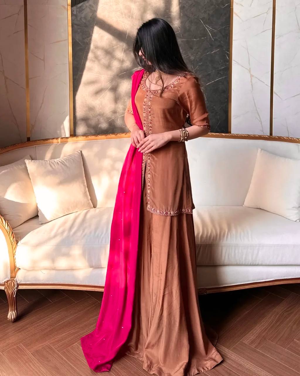 Woman in a brown saree with a pink blouse standing in a room with a white sofa and decorative wall.
