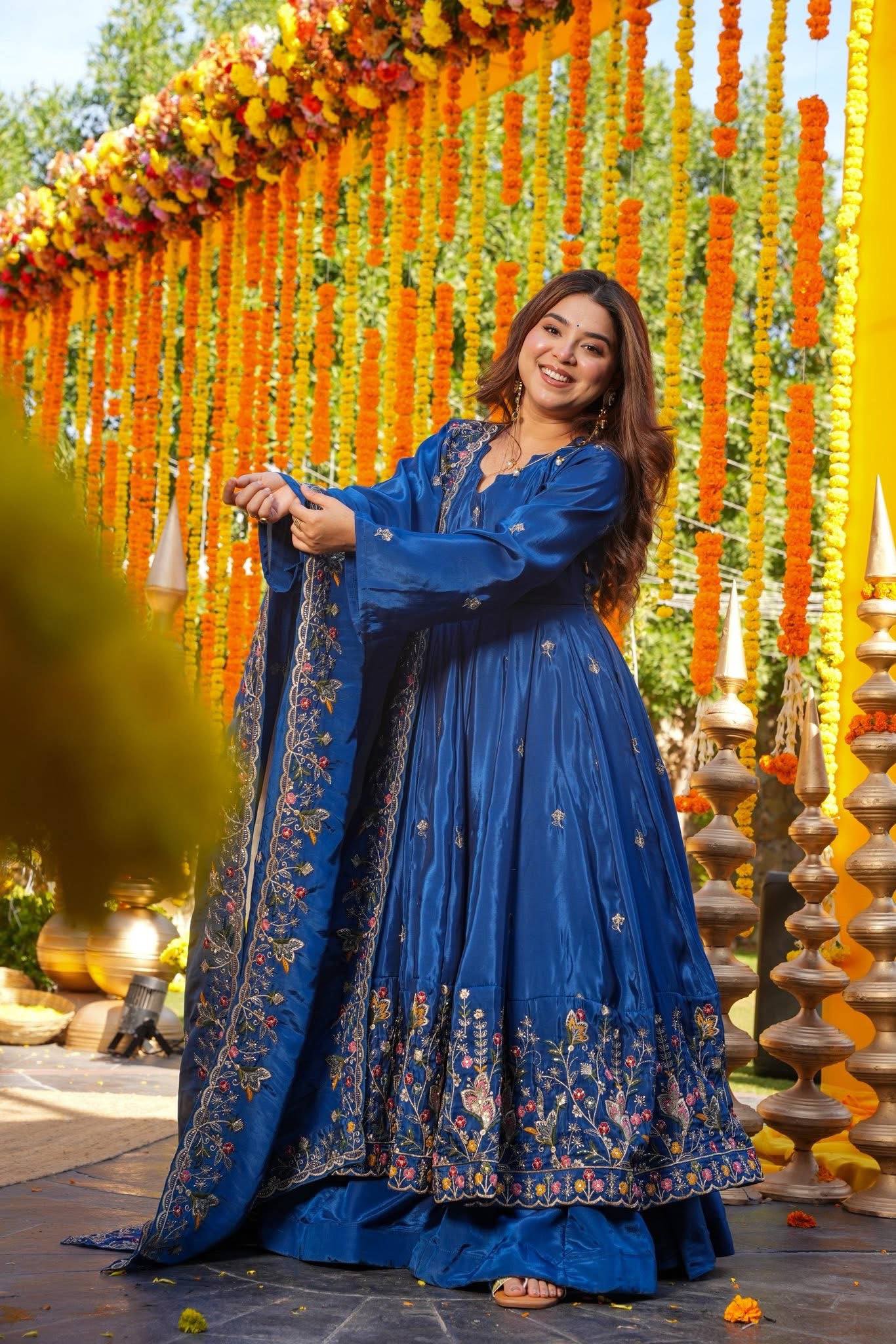 Woman in a blue traditional outfit standing in front of decorative floral arrangements.