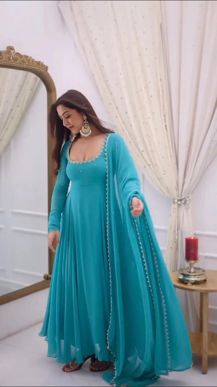 Woman in a turquoise dress with long sleeves and embellishments standing in a room with white curtains and a mirror.